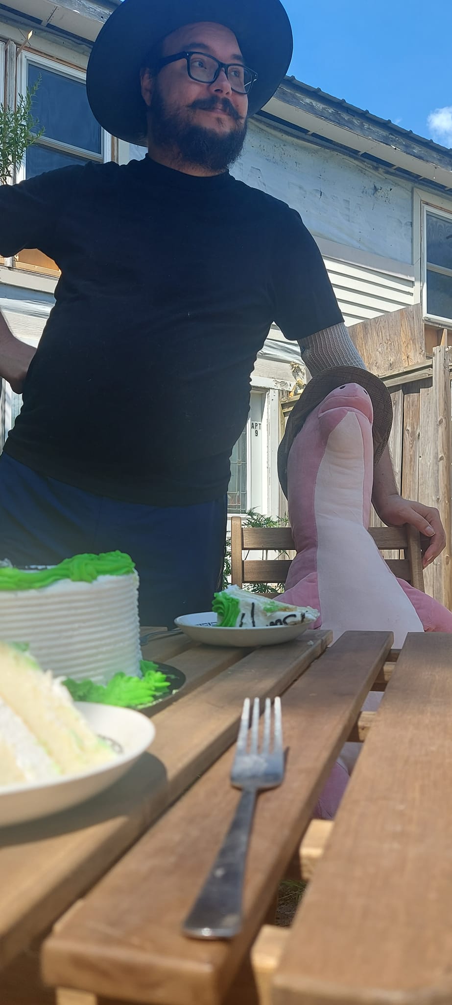 A man with facial hair and a felt sun hat has is standing with a pink dinosaur plush at a stuffed animal patio party